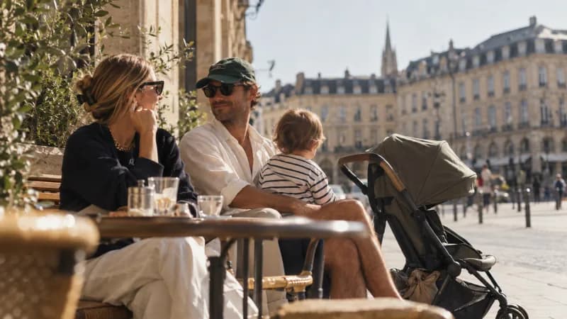 Un couple attablé à une terrasse de café ensoleillée avec un jeune enfant, une poussette compacte garée à côté de la table, dans une rue bordée d'immeubles anciens.